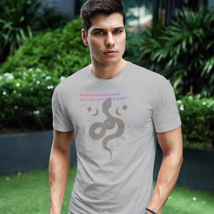 May include: A light gray t-shirt featuring a stylized snake design with text. The text reads "The urge to save humanity is almost always a false front for the urge to rule it." The shirt is worn by a person.