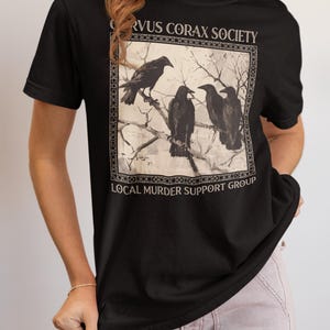 Op de afbeelding: Een zwart T-shirt met korte mouwen en een rechthoekige afbeelding van vier raven op kale boomtakken. De tekst CORVUS CORAX SOCIETY staat boven de afbeelding en LOCAL MURDER SUPPORT GROUP is in het wit onder de afbeelding gedrukt.
