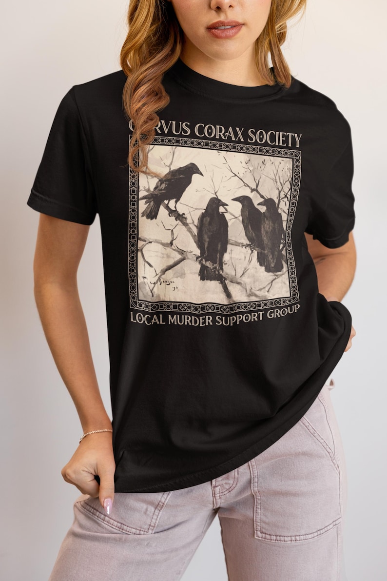Op de afbeelding: Een zwart T-shirt met korte mouwen en een rechthoekige afbeelding van vier raven op kale boomtakken. De tekst CORVUS CORAX SOCIETY staat boven de afbeelding en LOCAL MURDER SUPPORT GROUP is in het wit onder de afbeelding gedrukt.