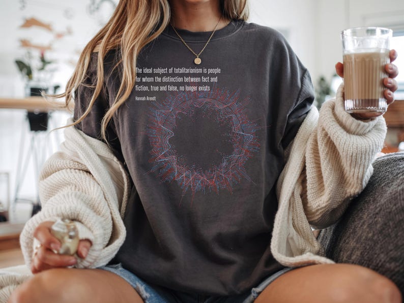 May include: A charcoal gray crewneck sweatshirt featuring a circular abstract graphic in red and blue lines. The text reads: The ideal subject of totalitarianism is people for whom the distinction between fact and fiction, true and false, no longer exists. Hannah Arendt.