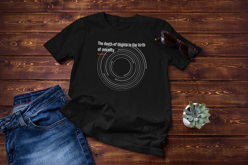 May include: Black t-shirt with the text "The death of dogma is the birth of morality" and a circular graphic. The shirt is paired with blue jeans and sunglasses. A small potted succulent is also visible.