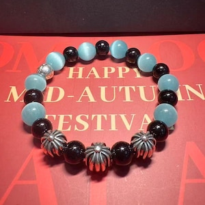 Könnte beinhalten: Ein Armband mit hellblauen und schwarzen Perlen, abwechselnd mit silberfarbenen Zierelementen. Das Armband liegt auf einer roten Oberfläche mit der Aufschrift "HAPPY MID-AUTUMN FESTIVAL".