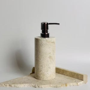 May include: A cylindrical soap dispenser made of beige marble with a polished chrome pump. The dispenser sits on a matching marble tray, creating a cohesive and elegant look. The design is minimalist and suitable for a bathroom or kitchen.