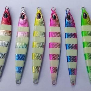May include: Seven colorful fishing lures with a metallic finish and horizontal stripes. Each lure has a different color scheme, including green, red, pink, yellow, and blue. The lures are designed for attracting fish.