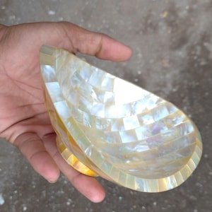 Pode incluir: Uma tigela decorativa em forma de gota, feita de pequenas telhas quadradas iridescentes. A tigela tem uma pequena base redonda e é segurada em uma mão. As telhas em mosaico criam um efeito perolado.