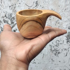May include: A small, handcrafted wooden cup with a curved handle. The cup is made of light-coloured wood, showcasing the natural grain. The handle extends from the side, creating a unique, ergonomic design. This rustic cup is perfect for enjoying coffee or tea.
