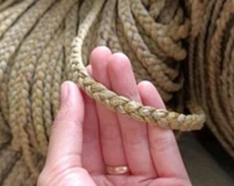 May include: Braided natural fiber rope, tan in color, held in a hand. The rope is thick and appears to be made of woven strands. The background shows more coiled rope. This rope could be used for crafting or decorative purposes.
