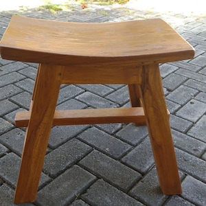 Puede incluir: Un pequeño taburete de madera con un asiento curvo y cuatro patas anguladas. El taburete es de madera marrón claro y tiene un diseño simple y minimalista. El asiento mide aproximadamente 30 cm de ancho y 25 cm de profundidad.