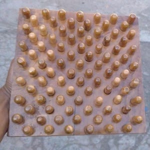 Op de afbeelding: Een vierkante houten massagetool met talrijke afgeronde houten pinnen. De pinnen zijn in een rasterpatroon gerangschikt en hebben een lichtere bruintint dan de basis. De tool wordt door een hand vastgehouden.