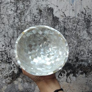 May include: A decorative bowl with a mosaic pattern made of iridescent white shell pieces. The bowl has a rounded shape and a smooth, reflective surface. The bowl is held in a hand against a textured gray background.