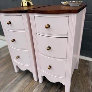 Op de afbeelding: Twee bijpassende roze nachtkastjes met gebogen lades en donkerbruine bladen. Elke tafel heeft drie lades met messing knoppen en decoratieve lijsten. Een gouden Eiffeltoren beeldje en een klein schaaltje staan erop.