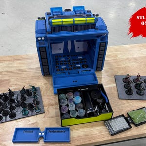 May include: A blue 3D printer case with yellow accents, open to reveal internal components. Several miniature figurines are displayed on gray platforms, along with paint bottles and tools. The text "STL FILES ONLY!" is visible.