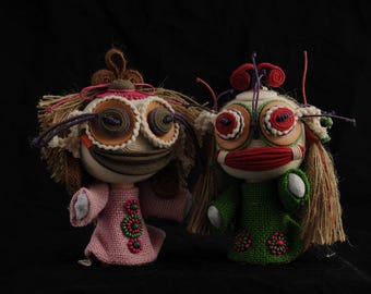 Puede incluir: Dos muñecas hechas a mano con rasgos caprichosos. Una muñeca viste un vestido rosa, la otra un vestido verde. Ambas tienen ojos grandes y redondos, pelo de hilo y detalles decorativos. Las muñecas están sobre un fondo negro.