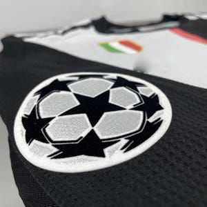 May include: Close-up of a black and white soccer jersey. The jersey features a large, embroidered soccer ball logo with a star design. The jersey has a mesh texture and a small emblem with green, white, and red stripes.