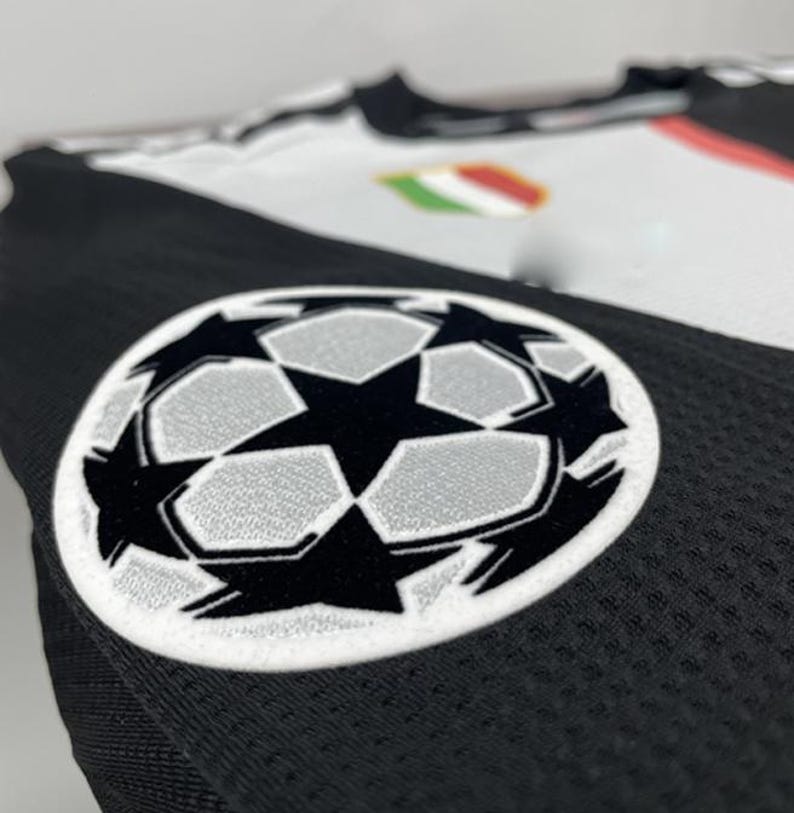 May include: Close-up of a black and white soccer jersey. The jersey features a large, embroidered soccer ball logo with a star design. The jersey has a mesh texture and a small emblem with green, white, and red stripes.