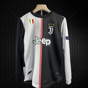 May include: A long-sleeved soccer jersey, split vertically with white and black panels. The white panel features the "Jeep" logo, while the black panel has the "Juventus" logo. A red stripe runs down the center. The jersey has a round neck and a small Italian flag patch.
