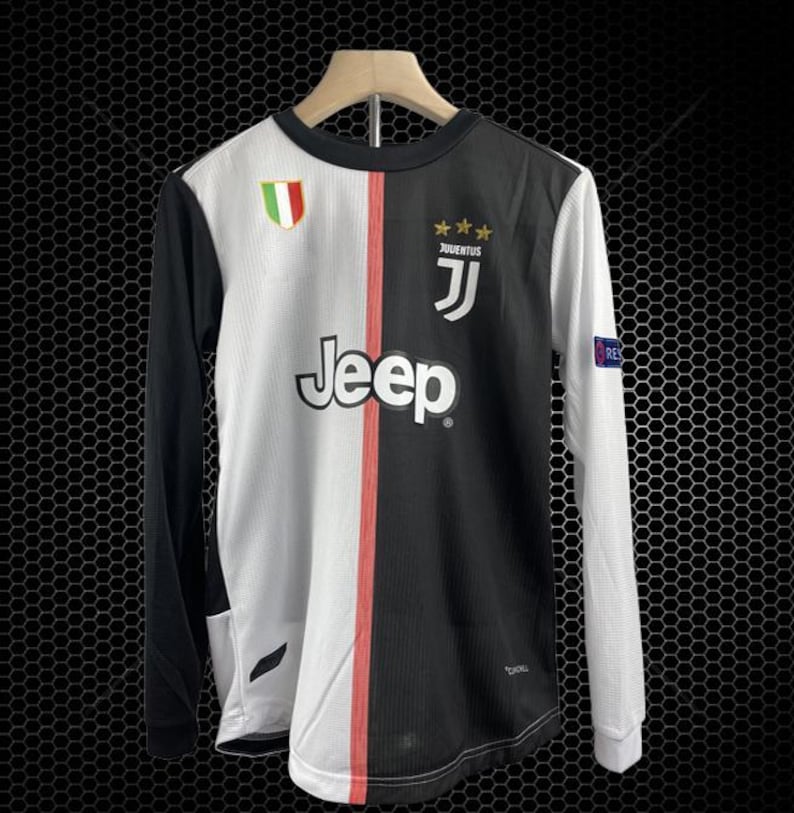 May include: A long-sleeved soccer jersey, split vertically with white and black panels. The white panel features the "Jeep" logo, while the black panel has the "Juventus" logo. A red stripe runs down the center. The jersey has a round neck and a small Italian flag patch.