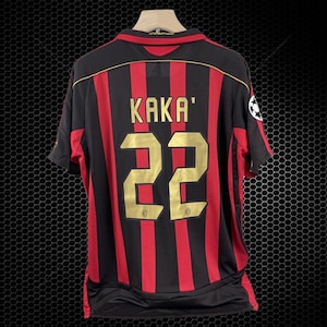 Puede incluir: Camiseta de fútbol a rayas negras y rojas con el nombre "KAKA'" y el número "22" en letras doradas. La camiseta tiene cuello redondo y mangas cortas. Un logotipo de la Liga de Campeones es visible en la manga derecha.