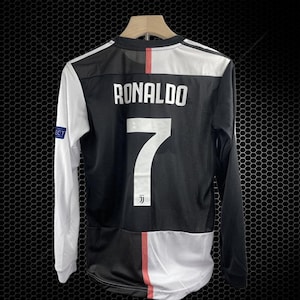 Puede incluir: Camiseta de fútbol de manga larga en blanco y negro con el nombre "RONALDO" y el número "7" en la espalda. La camiseta tiene una raya roja en la espalda y mangas blancas. Cuelga de una percha de madera.