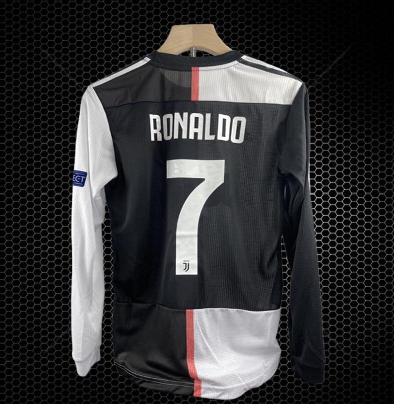 May include: Black and white long-sleeved soccer jersey with the name "RONALDO" and the number "7" on the back. The jersey has a red stripe down the back and white sleeves. The jersey is hanging on a wooden hanger.