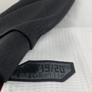 May include: Close-up of a black and white athletic jersey. The jersey features a black collar and sleeve detail. A black patch with the text "19/20 AUTHENTIC" is sewn onto the jersey.