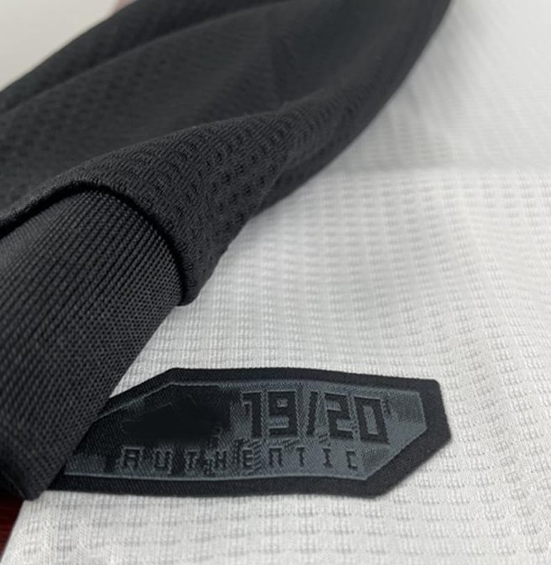 May include: Close-up of a black and white athletic jersey. The jersey features a black collar and sleeve detail. A black patch with the text "19/20 AUTHENTIC" is sewn onto the jersey.
