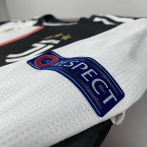 May include: Close-up of a white and black soccer jersey. The jersey features the word "RESPECT" on the sleeve, with a blue and red emblem. The jersey has a textured pattern and the Juventus logo is visible.