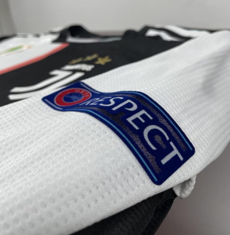 May include: Close-up of a white and black soccer jersey. The jersey features the word "RESPECT" on the sleeve, with a blue and red emblem. The jersey has a textured pattern and the Juventus logo is visible.