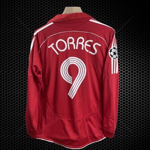 Può includere: Maglia da calcio rossa a maniche lunghe con dettagli bianchi. La maglia presenta il nome "TORRES" e il numero "9" in bianco sul retro. Una grafica di pallone da calcio è sulla manica.