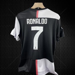 Puede incluir: Camiseta de fútbol en blanco y negro con el nombre "RONALDO" y el número "7" en la espalda. La camiseta tiene una franja roja y un logotipo del equipo. Cuelga de una percha de madera.