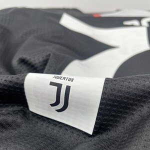 May include: Close-up of a black and white Juventus soccer jersey. The jersey features the Juventus logo and the word "JUVENTUS" on a white label. The fabric has a textured, mesh-like appearance.