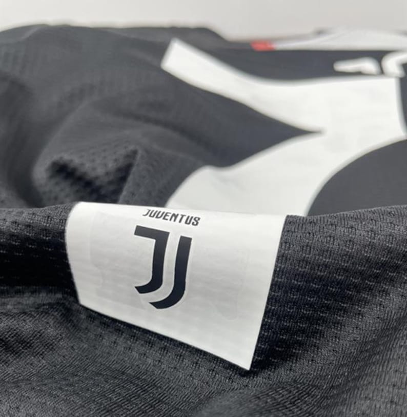 May include: Close-up of a black and white Juventus soccer jersey. The jersey features the Juventus logo and the word "JUVENTUS" on a white label. The fabric has a textured, mesh-like appearance.