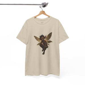 May include: A beige t-shirt displaying a seated fairy with brown skin, spiky hair, and golden, glittery wings. The fairy is the central focus, set against the neutral backdrop of the shirt. The t-shirt is suspended from a wooden hanger.