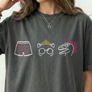 May include: Dark gray Comfort Colors t-shirt with three whimsical illustrations: boxer shorts with heart shapes, a cat with glasses and a crown, and a dinosaur. The word "PEPPER" is printed on the bottom right.