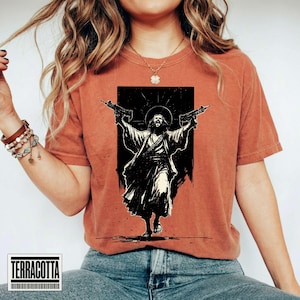 May include: A rust-colored short-sleeve t-shirt featuring a black graphic print of a figure with long hair and a robe, holding two firearms aloft with a halo effect behind the head. The design is centered on the chest of the garment.