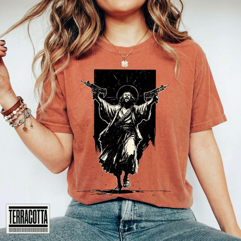 May include: A rust-colored short-sleeve t-shirt featuring a black graphic print of a figure with long hair and a robe, holding two firearms aloft with a halo effect behind the head. The design is centered on the chest of the garment.