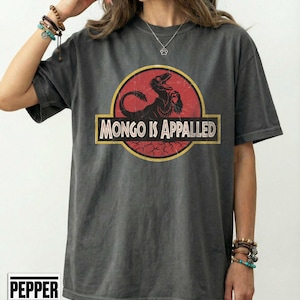 May include: Dark gray t-shirt featuring a dinosaur graphic inside a red circle with the text "MONGO IS APPALLED" in a yellow rectangle. The shirt has a vintage, worn look.