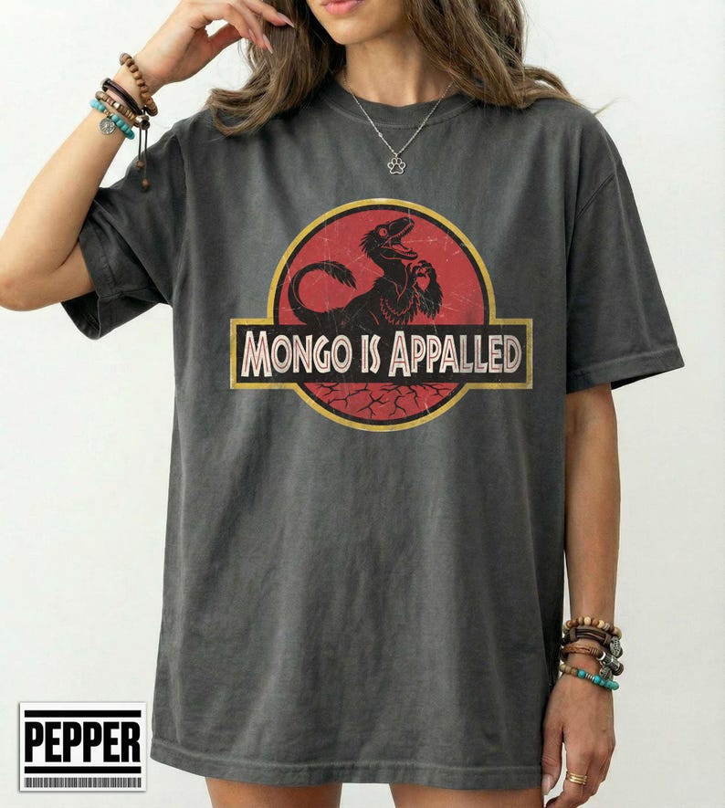 Może przedstawiać: Ciemnoszary t-shirt z kr&oacute;tkim rękawem z okrągłą grafiką na czerwonym tle, przedstawiającą sylwetkę czarnego dinozaura. Napis "MONGO IS APPALED" jest nadrukowany pogrubioną, jasną czcionką na środku okrągłego wzoru.