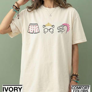 May include: An ivory short-sleeve t-shirt featuring a graphic print of three icons: a pair of boxer shorts with red hearts, a cat face wearing a gold crown and glasses, and a dinosaur head with a pink crest. The shirt has a crew neckline.