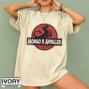 Może przedstawiać: T-shirt z kr&oacute;tkim rękawem w kolorze kości słoniowej z okrągłą grafiką na czerwonym tle. Grafika przedstawia czarną sylwetkę dinozaura oraz napis "MONGO IS APPALLED" pogrubioną czcionką szeryfową. Koszulka posiada okrągły dekolt.