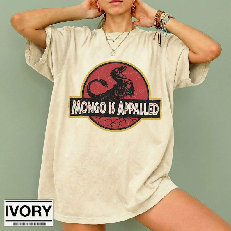 Może przedstawiać: T-shirt z kr&oacute;tkim rękawem w kolorze kości słoniowej z okrągłą grafiką na czerwonym tle. Grafika przedstawia czarną sylwetkę dinozaura oraz napis "MONGO IS APPALLED" pogrubioną czcionką szeryfową. Koszulka posiada okrągły dekolt.