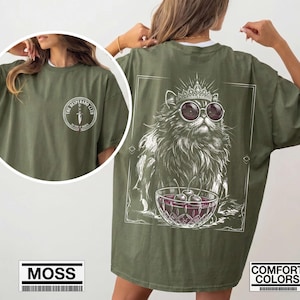 May include: An olive green t-shirt featuring a white graphic on the back of a cat wearing a crown and round sunglasses, sitting behind a bowl of cherries. The front chest area displays a circular logo with a dagger and the text THE DESPERADO CLUB.