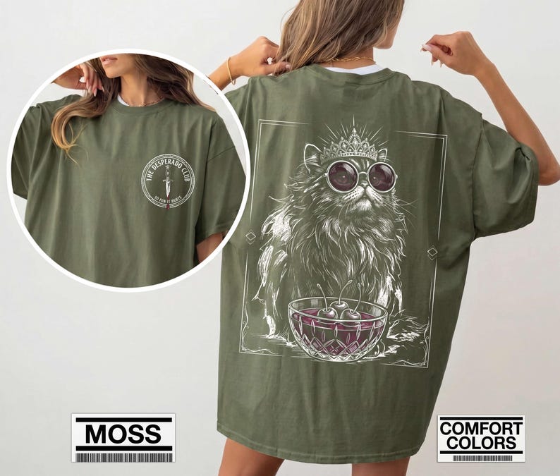 May include: An olive green t-shirt featuring a white graphic on the back of a cat wearing a crown and round sunglasses, sitting behind a bowl of cherries. The front chest area displays a circular logo with a dagger and the text THE DESPERADO CLUB.