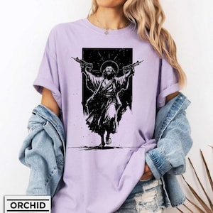 May include: A light purple t-shirt featuring a black graphic print of a figure with a halo holding two firearms aloft. The design is centered on the chest, showcasing a high-contrast illustration style with detailed line work and a dark rectangular background.