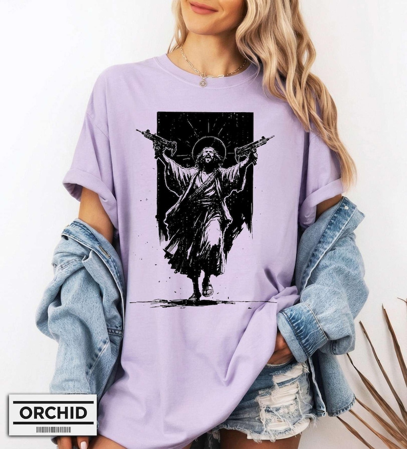 May include: A light purple t-shirt featuring a black graphic print of a figure with a halo holding two firearms aloft. The design is centered on the chest, showcasing a high-contrast illustration style with detailed line work and a dark rectangular background.