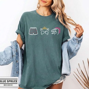 May include: A dark teal short-sleeve t-shirt featuring a graphic print of white boxer shorts with red hearts, a white cat outline wearing a gold crown and glasses, and a white dinosaur outline with a pink crest. The shirt has a crew neckline.