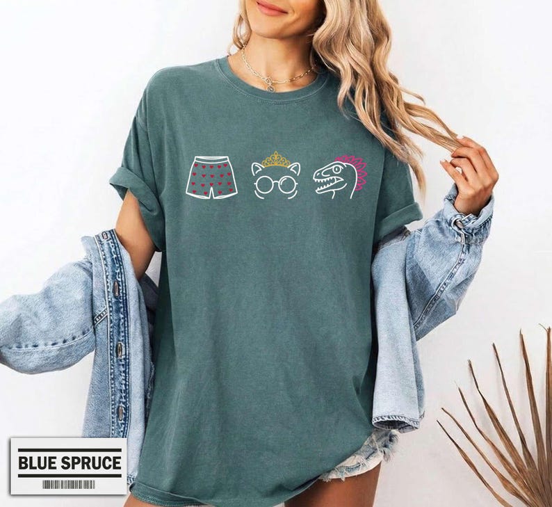 May include: A dark teal short-sleeve t-shirt featuring a graphic print of white boxer shorts with red hearts, a white cat outline wearing a gold crown and glasses, and a white dinosaur outline with a pink crest. The shirt has a crew neckline.