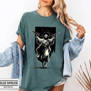 May include: A blue-green crew neck t-shirt featuring a black graphic print of a bearded figure in robes holding two rifles with arms raised. The shirt has a relaxed fit and short sleeves, paired with a light blue denim jacket draped over the shoulders.