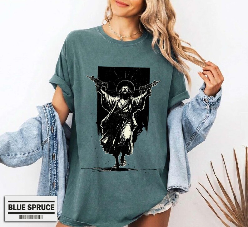 May include: A blue-green crew neck t-shirt featuring a black graphic print of a bearded figure in robes holding two rifles with arms raised. The shirt has a relaxed fit and short sleeves, paired with a light blue denim jacket draped over the shoulders.