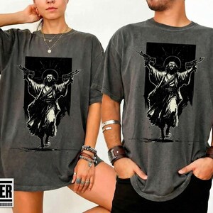 May include: A charcoal gray short-sleeve t-shirt featuring a black and white graphic print of a figure in robes with a halo, holding a firearm in each outstretched hand. The design is centered on the chest of the garment.